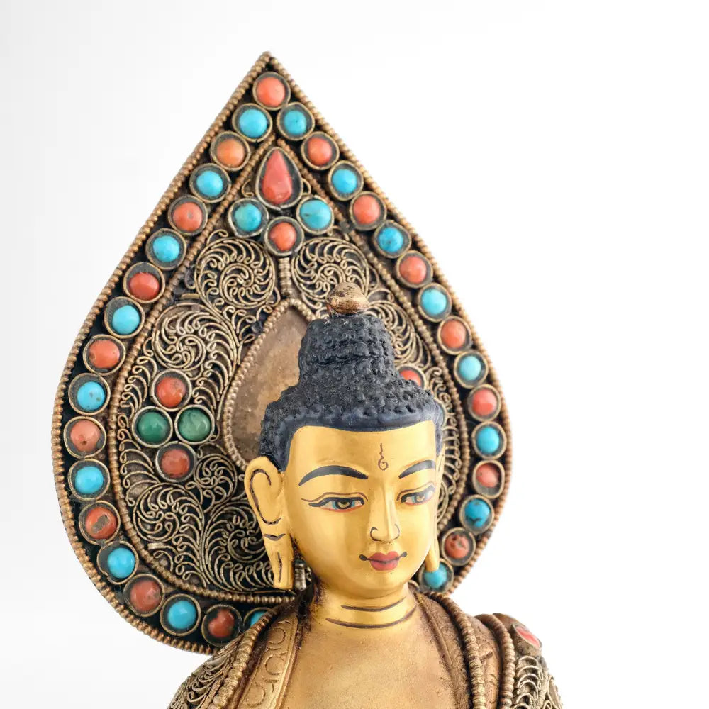 Gesicht der Shakyamuni Buddha Figur. Vor dem tropfenförmigen Hintergrund, Nimbus. Der Hintergrund ist besetzt mit Türkis und Korallensteinen. Und feinen, versilberten Filigran Ornamenten in Spiralform.
