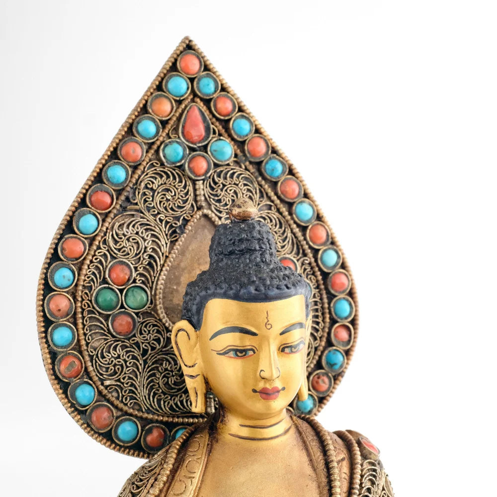 Gesicht der Shakyamuni Buddha Figur. Vor dem tropfenförmigen Hintergrund, Nimbus. Der Hintergrund ist besetzt mit Türkis und Korallensteinen. Und feinen, versilberten Filigran Ornamenten in Spiralform.