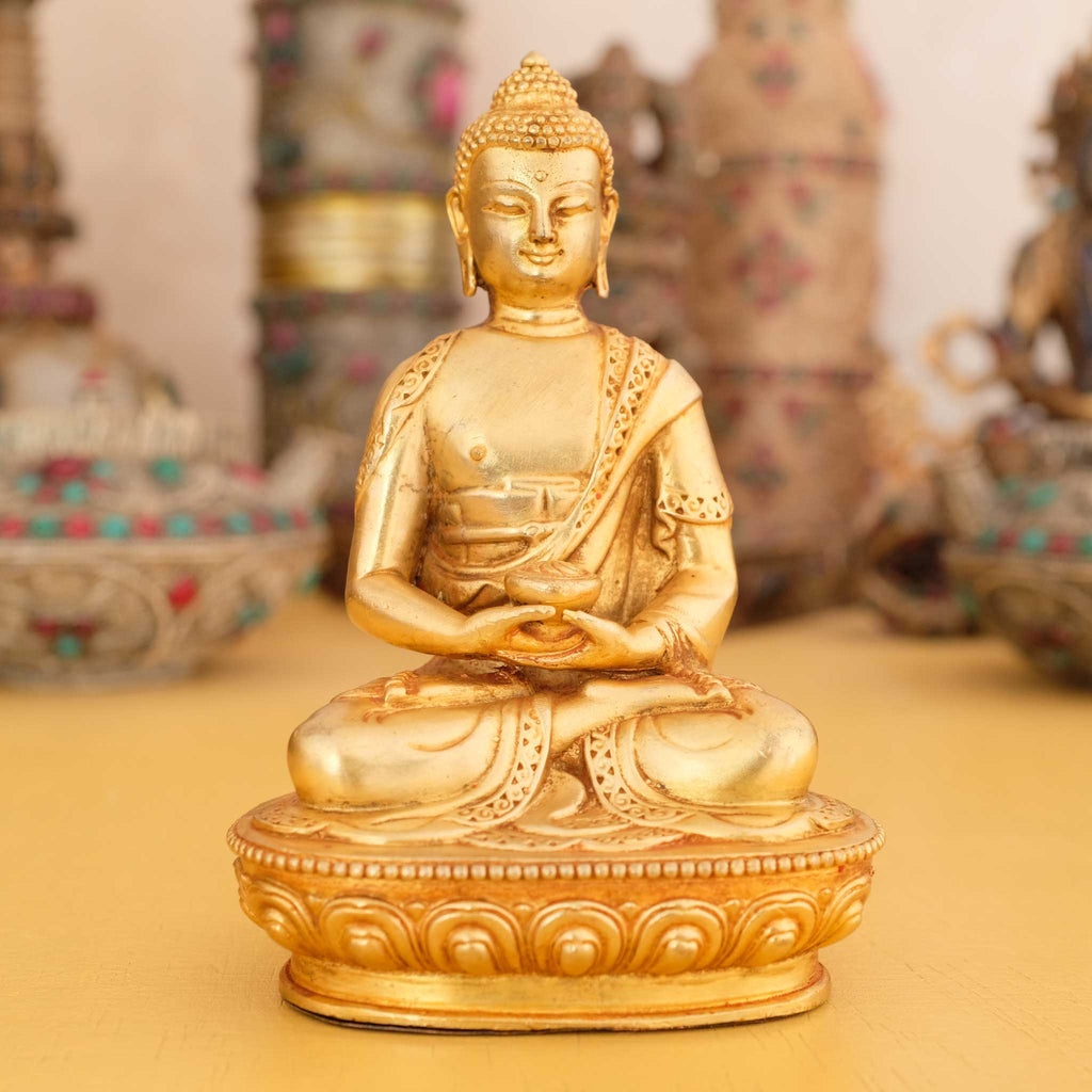 Kleine vergoldete Amitabha Buddha Statue, im Hintergrund rituelle Gegenstände des Vajrayana-Buddhismus in einer sanft beleuchteten Werkstattumgebung.