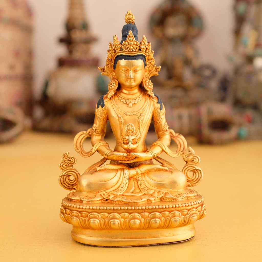 Kleine vergoldete Amitayus Buddha Statue, im Hintergrund rituelle Gegenstände des Vajrayana-Buddhismus in einer sanft beleuchteten Werkstattumgebung.