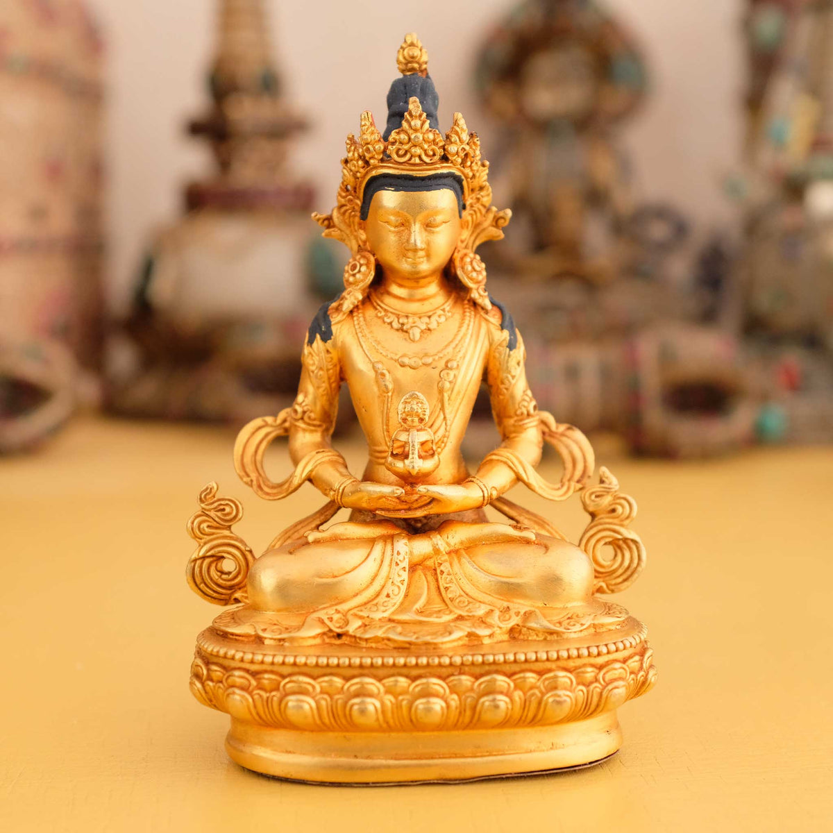 Kleine vergoldete Amitayus Buddha Statue, im Hintergrund rituelle Gegenstände des Vajrayana-Buddhismus in einer sanft beleuchteten Werkstattumgebung.