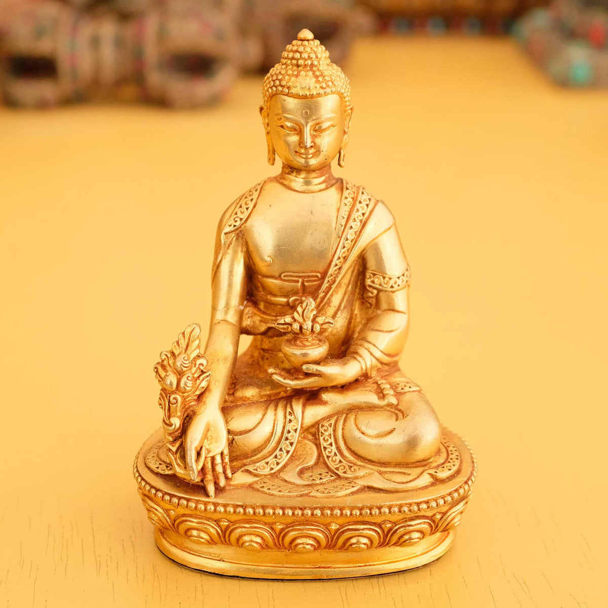 Kleine vergoldete Medizin Buddha Statue im Hintergrund rituelle Gegenstände des Vajrayana-Buddhismus in einer sanft beleuchteten Werkstattumgebung.