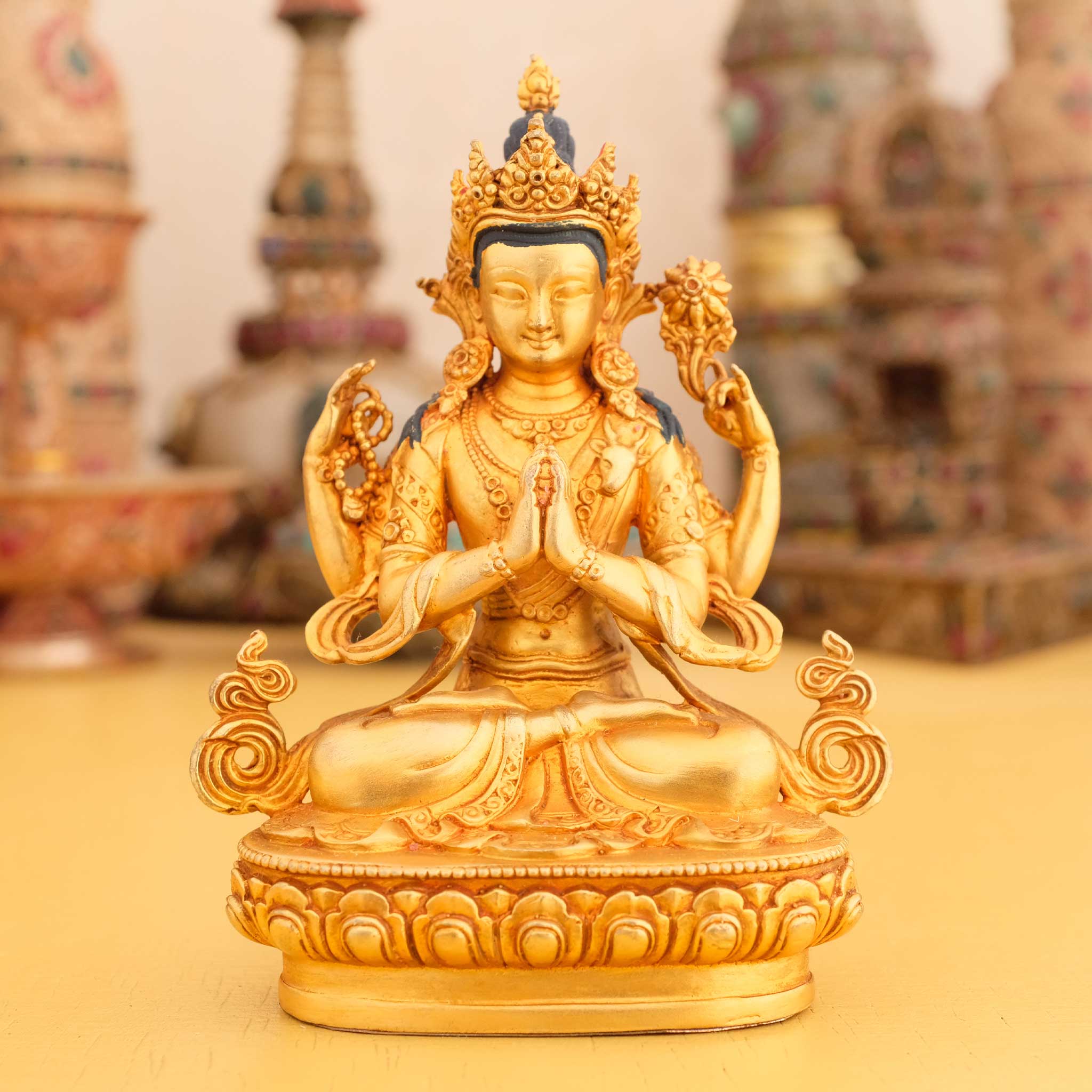 Kleine vergoldete Avalokiteshvara Statue, Chenrezig, im Hintergrund rituelle Gegenstände des Vajrayana-Buddhismus in einer sanft beleuchteten Werkstattumgebung.