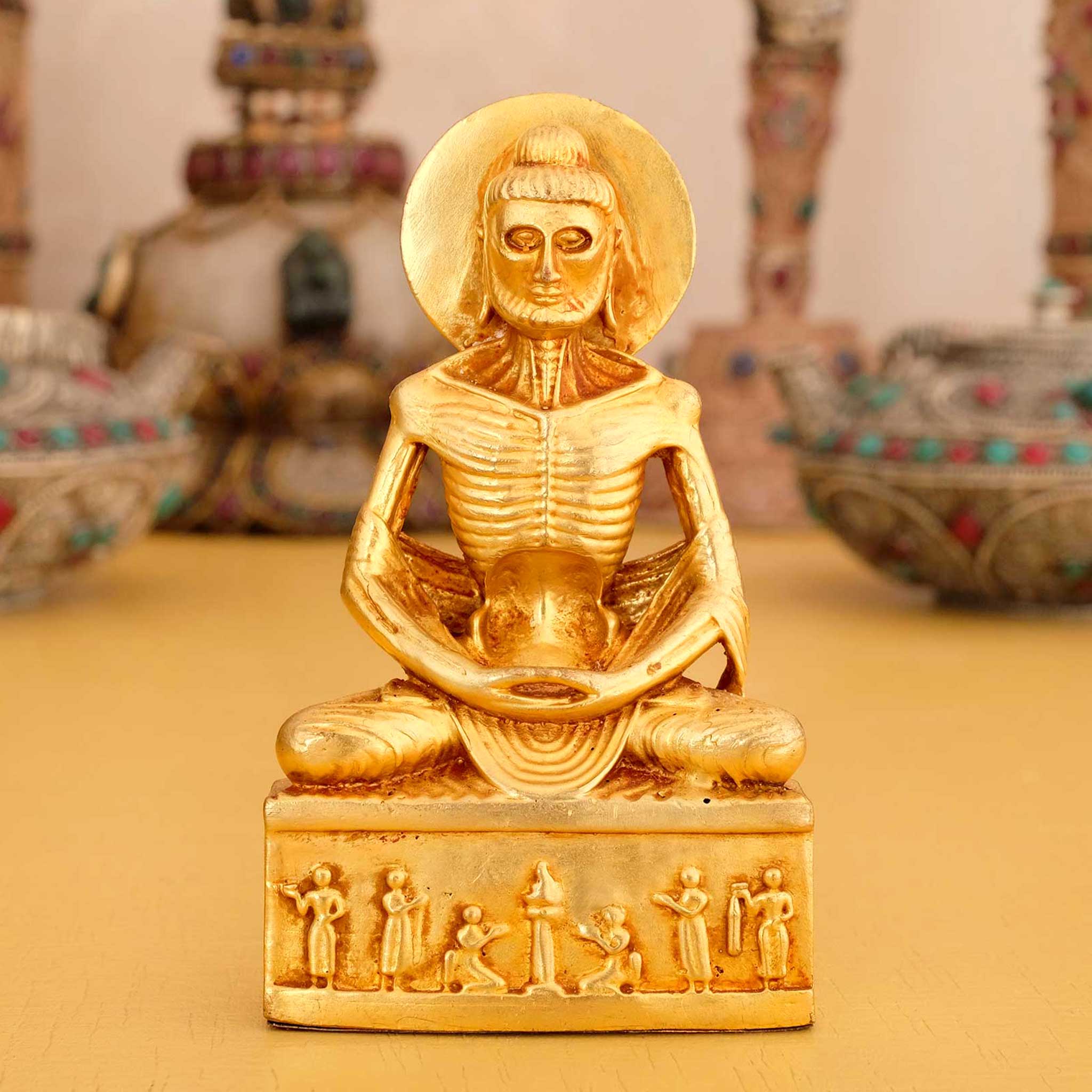 Kleine vergoldete Askese Buddha Statue, im Hintergrund rituelle Gegenstände des Vajrayana-Buddhismus in einer sanft beleuchteten Werkstattumgebung.
