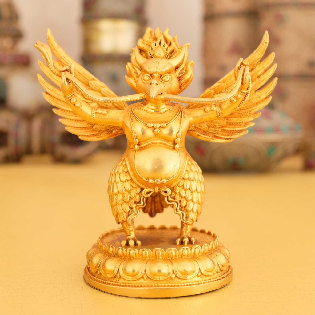 Kleine vergoldete Garuda Statue, im Hintergrund rituelle Gegenstände des Vajrayana-Buddhismus in einer sanft beleuchteten Werkstattumgebung.