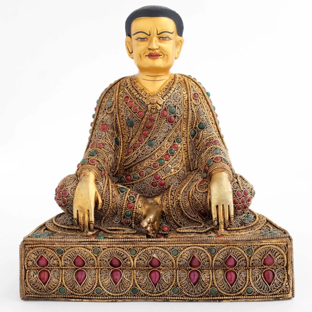Guru Marpa sitzt auf einem Lotus Sockel, seine Beine sind in einer locker verschränkten Haltung, seine Hände hängen über die Knie und berühren den Boden. Seine Robe und der Sockel sind reich verziert.