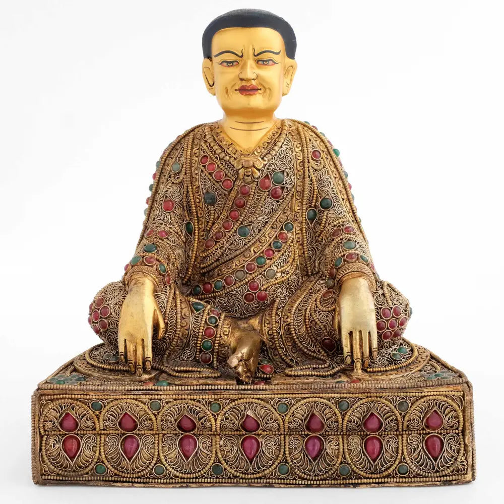 Guru Marpa sitzt auf einem Lotus Sockel, seine Beine sind in einer locker verschränkten Haltung, seine Hände hängen über die Knie und berühren den Boden. Seine Robe und der Sockel sind reich verziert.