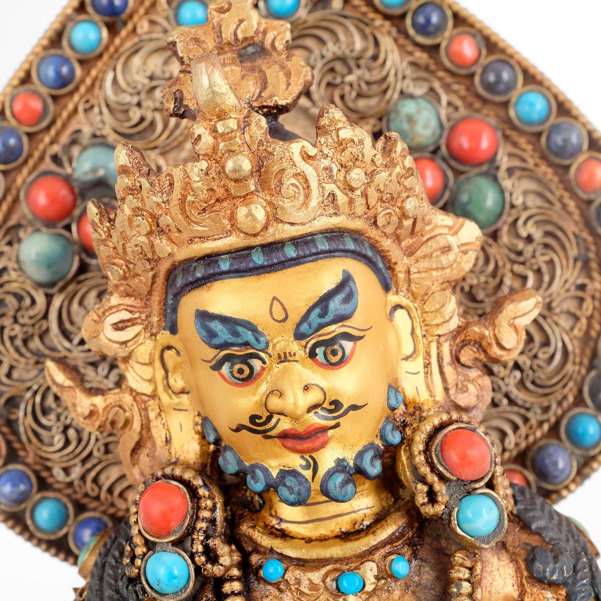 Detailaufnahme Jambhala Statue. Das Gesicht ist in Gold gefasst. Dahinter eine tropfenförmige Aureole die mit Filigran gestaltet ist und mit farbigen Steinen verziert.