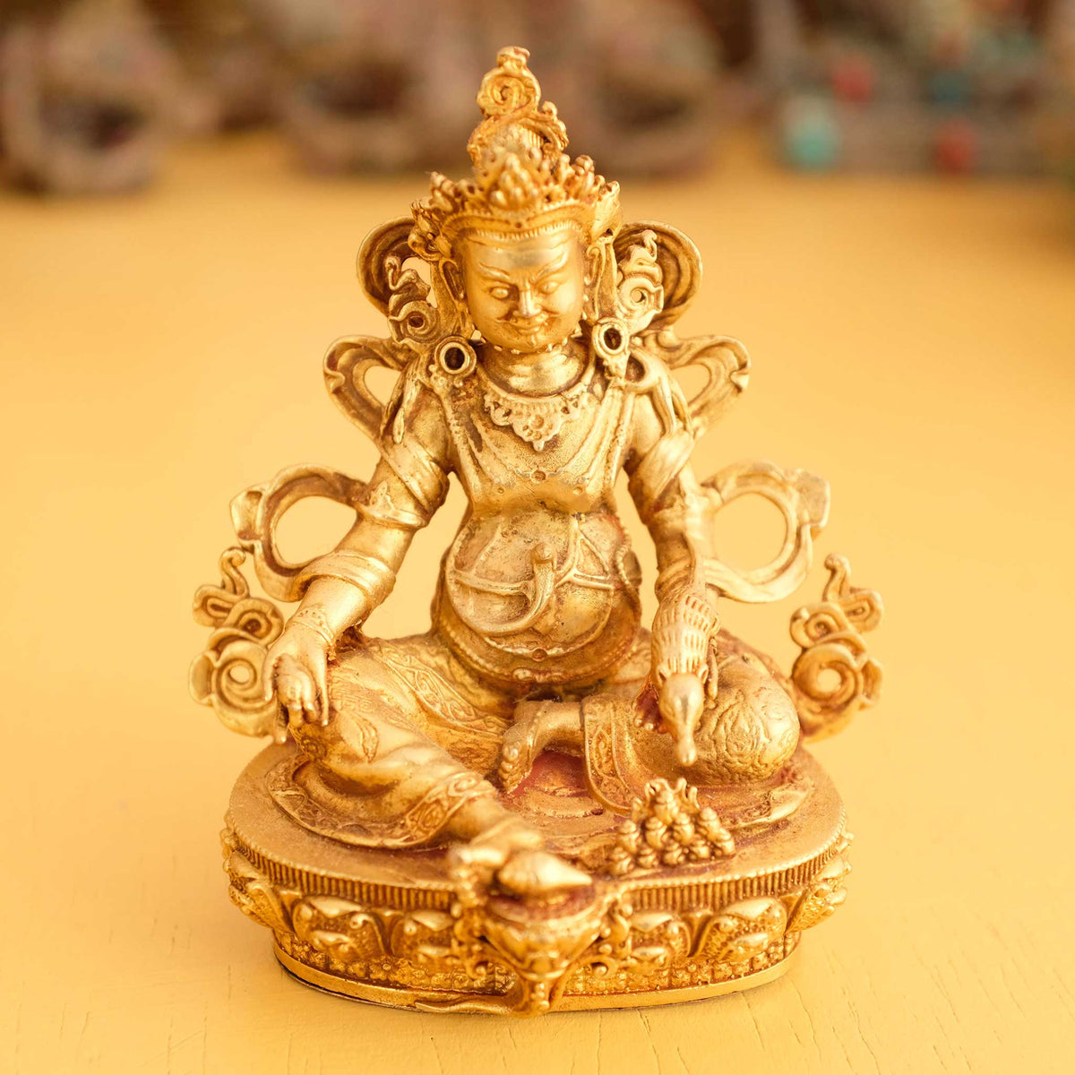 Kleine vergoldete Jambhala Statue, im Hintergrund rituelle Gegenstände des Vajrayana-Buddhismus in einer sanft beleuchteten Werkstattumgebung.