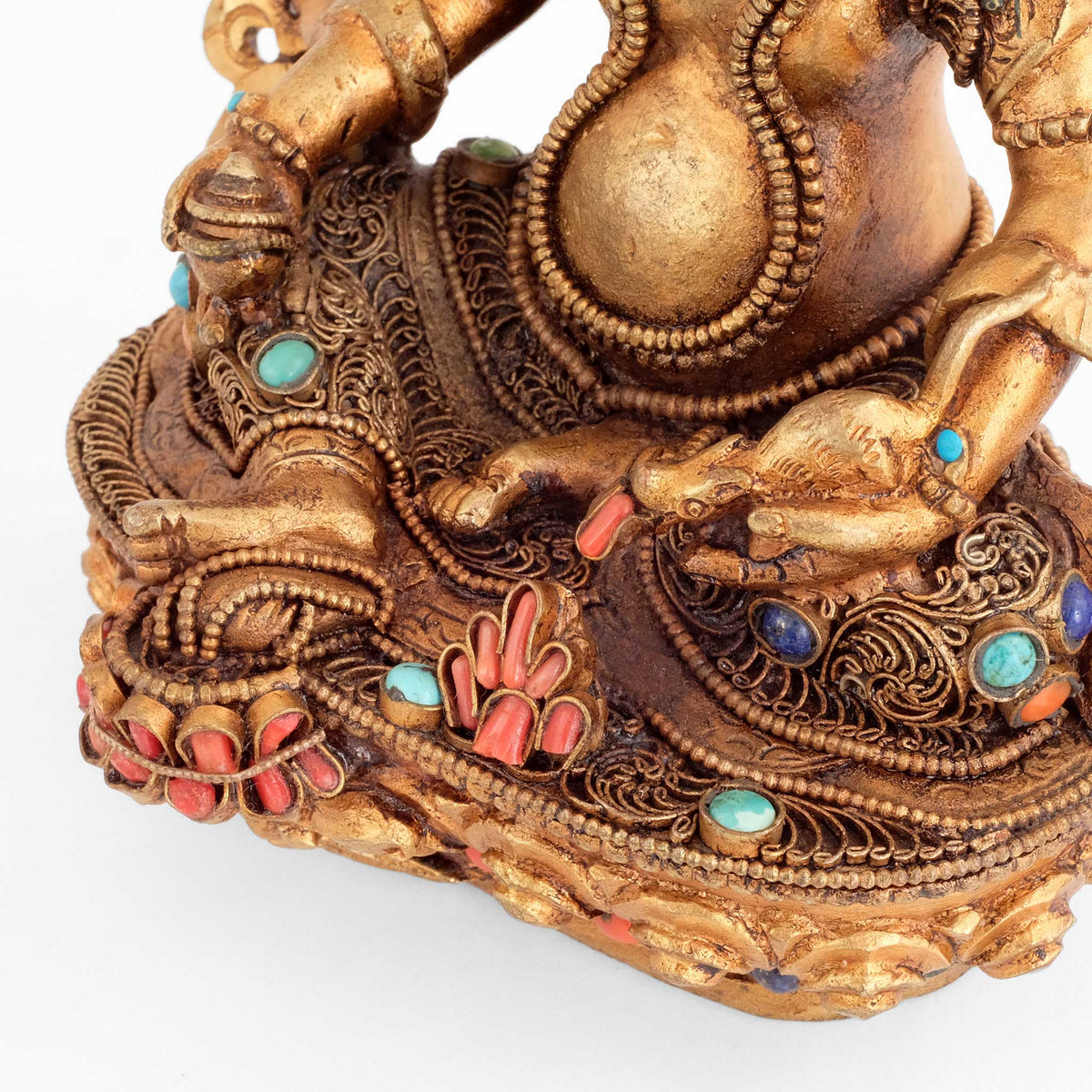 Jambhala Statue,  Aufsicht  auf die Beine inlalit asana, das Gewand mit Filigran gestaltet und mit farbigen Steinen besetzt. In seiner linken Hand hält er ein Mungo, Nakula das eine Zunge mit eingesetzter Koralle herausstreckt. Er spukt Juwelen die als Haufen am vorderen Rand des Lotussockels zu sehen sind.