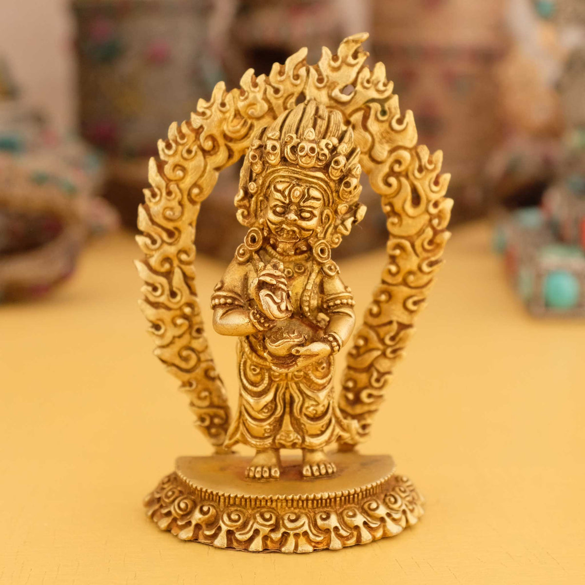 Kleine vergoldete Mahakala Statue, im Hintergrund rituelle Gegenstände des Vajrayana-Buddhismus in einer sanft beleuchteten Werkstattumgebung.