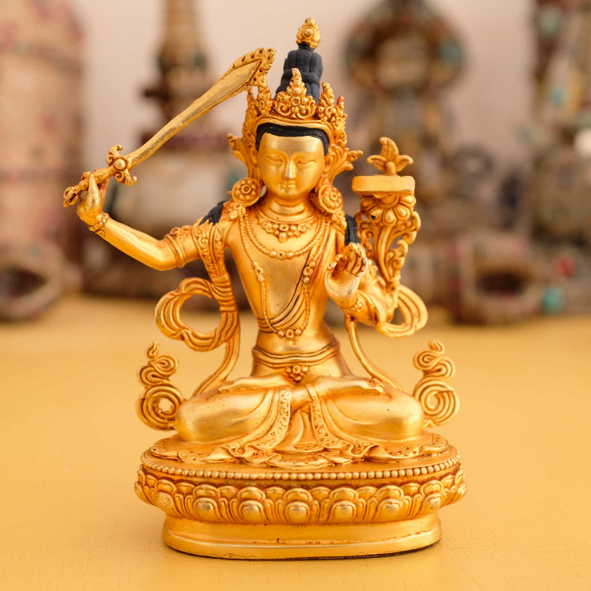 Kleine vergoldete Manjushri Statue, im Hintergrund rituelle Gegenstände des Vajrayana-Buddhismus in einer sanft beleuchteten Werkstattumgebung.