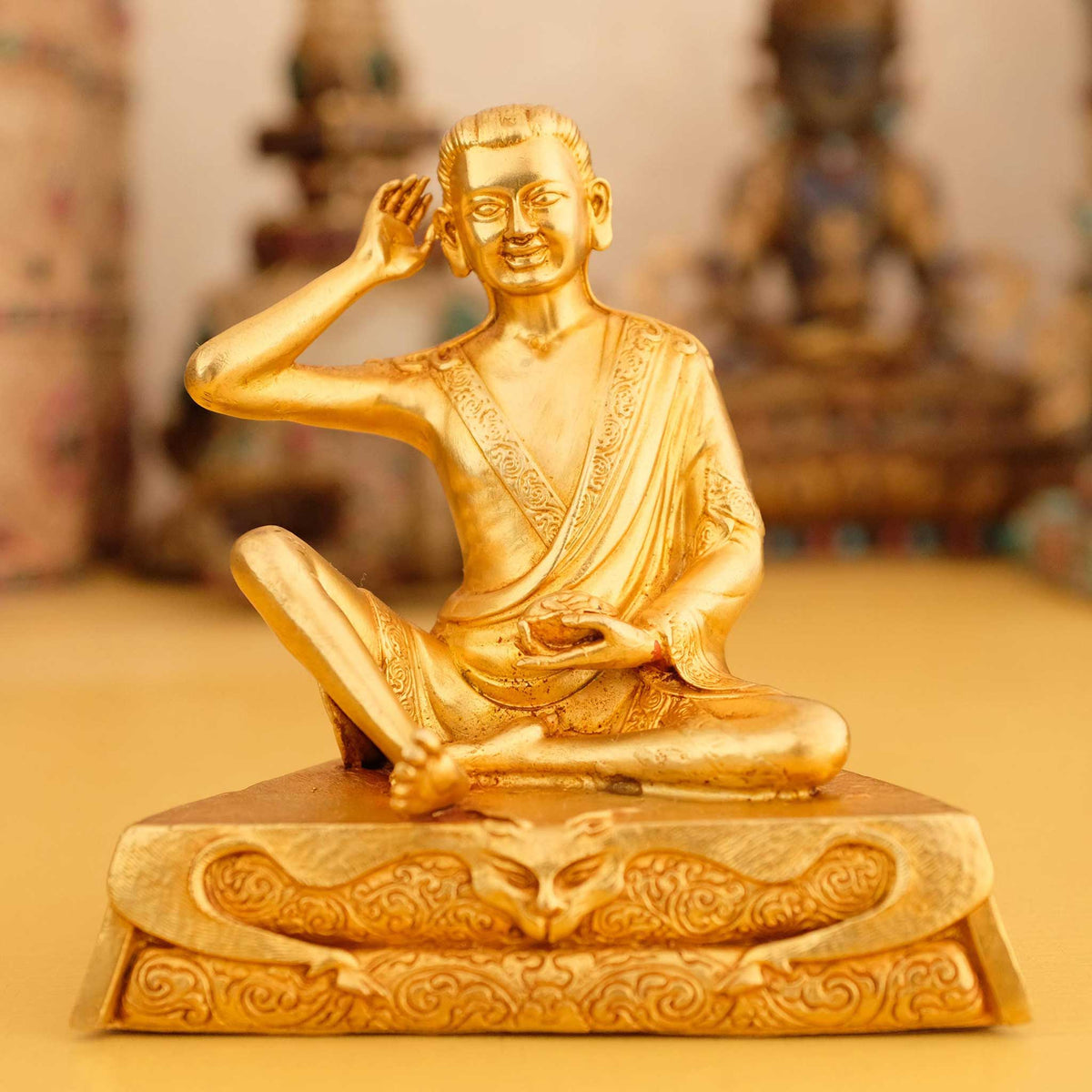 Kleine vergoldete Jetsun Milarepa Figur, im Hintergrund rituelle Gegenstände des Vajrayana-Buddhismus in einer sanft beleuchteten Werkstattumgebung.