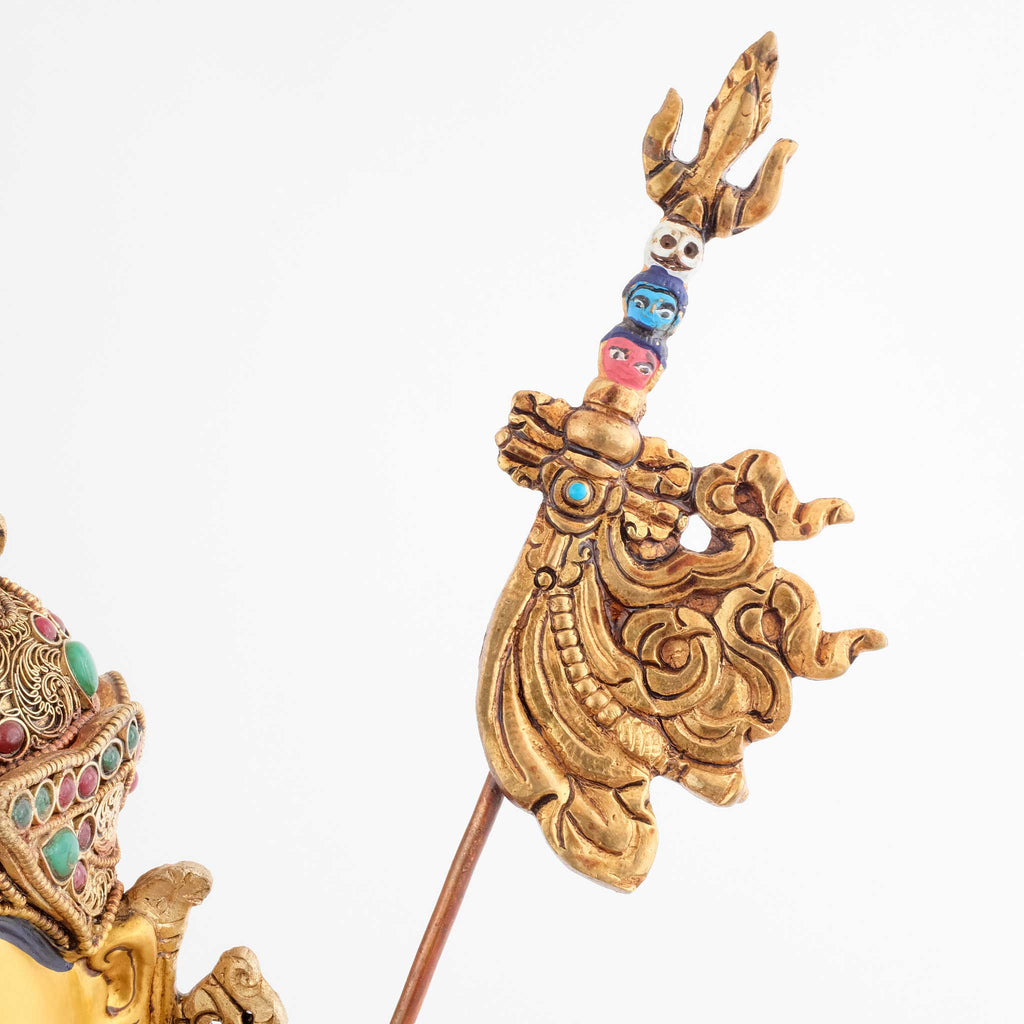 Der tantrische Stab Khatvanga, den Padmasambhava in seinem linken Arm hält, symbolisiert die weibliche Gefährtin als die Essenz der Weisheit. Er hat einen Dreizack an der Spitze, gefolgt von drei aufgespießten Köpfen in den Farben Weiß, Blau und Rot.