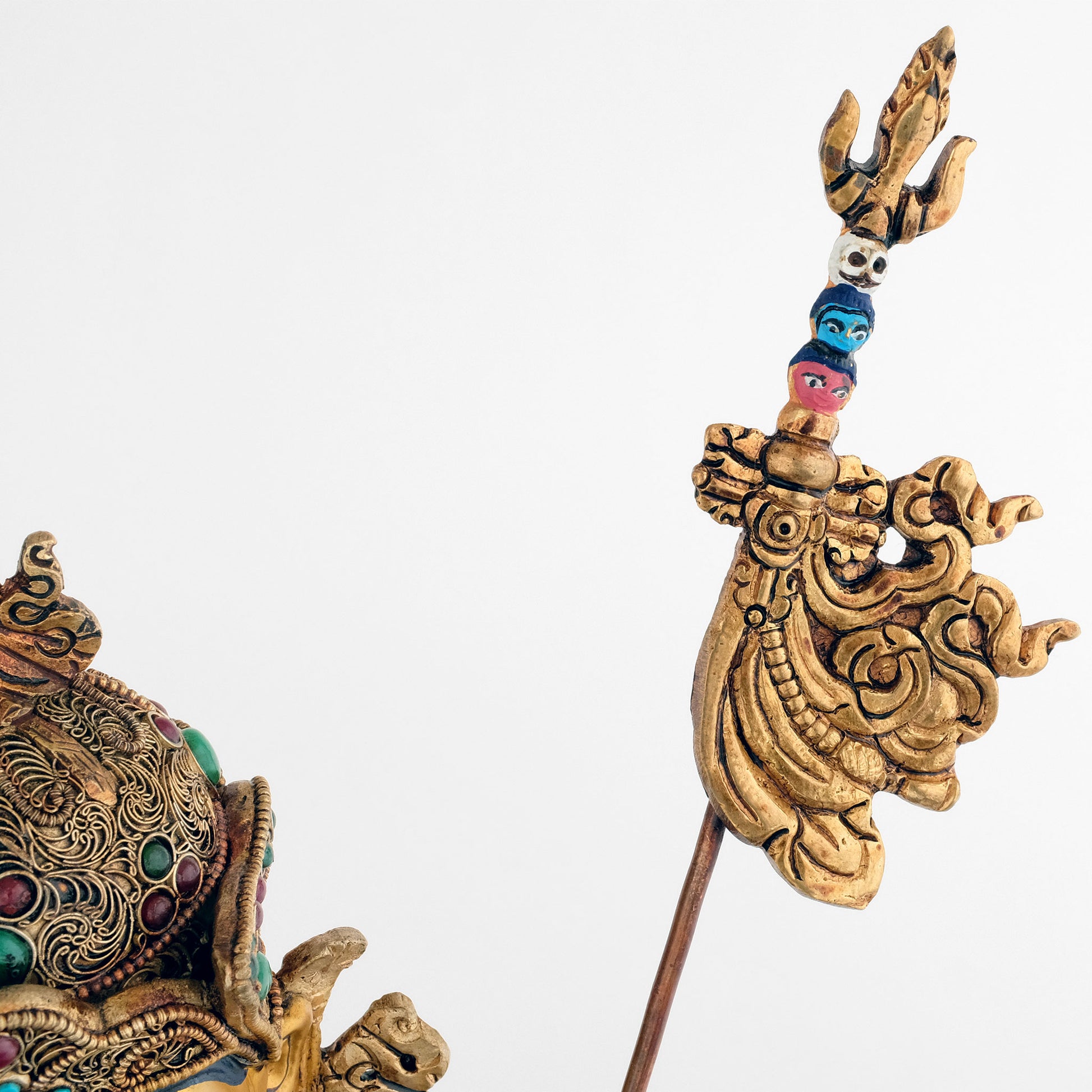 Der tantrische Stab Khatvanga, den Padmasambhava in seinem linken Arm hält, symbolisiert die weibliche Gefährtin als die Essenz der Weisheit. Er hat einen Dreizack an der Spitze, gefolgt von drei aufgespießten Köpfen in den Farben Weiß, Blau und Rot.