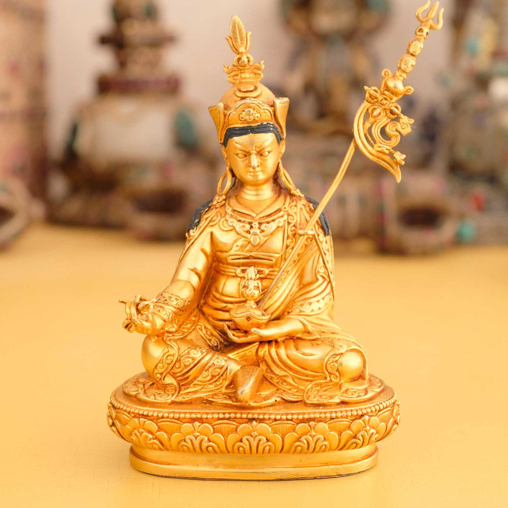 Kleine vergoldete Padmasambhava Statue, Guru Rinpoche, im Hintergrund rituelle Gegenstände des Vajrayana-Buddhismus in einer sanft beleuchteten Werkstattumgebung.