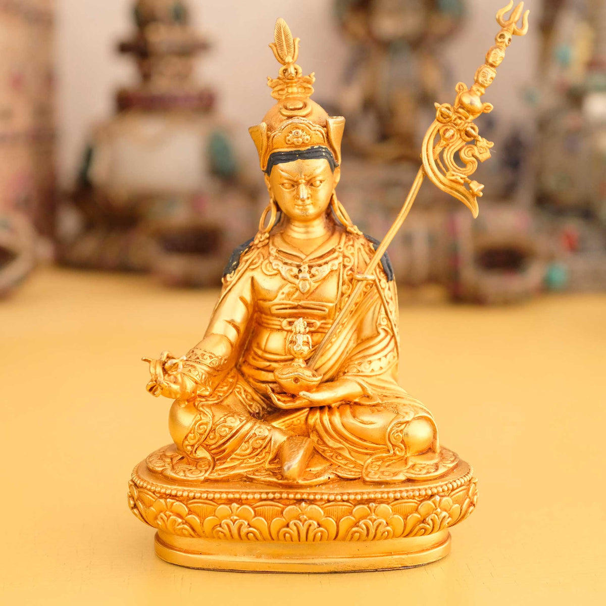 Kleine vergoldete Padmasambhava Statue, Guru Rinpoche, im Hintergrund rituelle Gegenstände des Vajrayana-Buddhismus in einer sanft beleuchteten Werkstattumgebung.