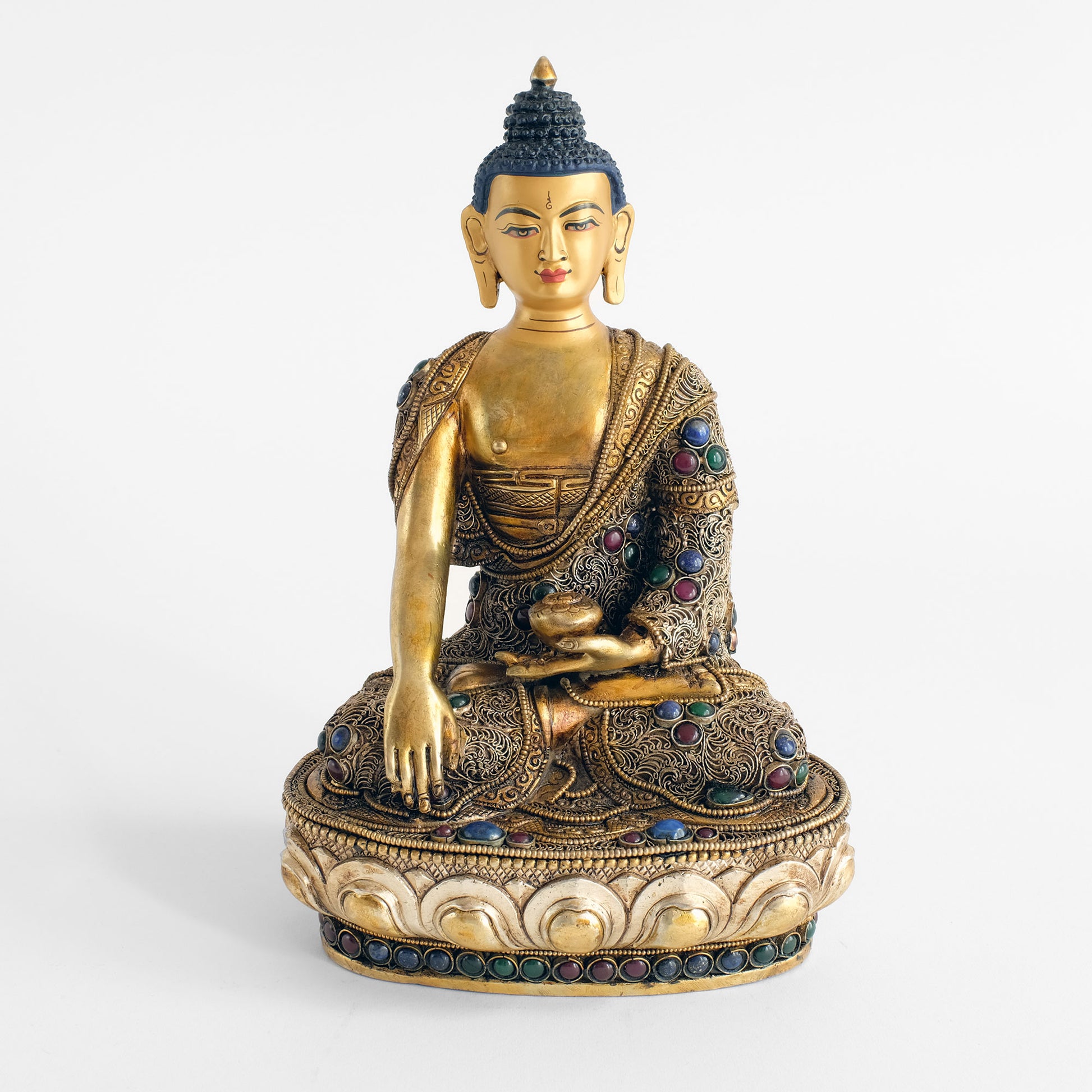 Buddha Shakyamuni Statue, sitzt in der Lotus Haltung, Padma Asana. Sein klösterliches Gewand ist mit feinem Filigran verziert, das in spiralförmigen Rosetten aufgelegt ist. Die Statue ist mit Dunkelroten, Dunkelblauen und Dunkelgrünen Steinen akzentuiert.