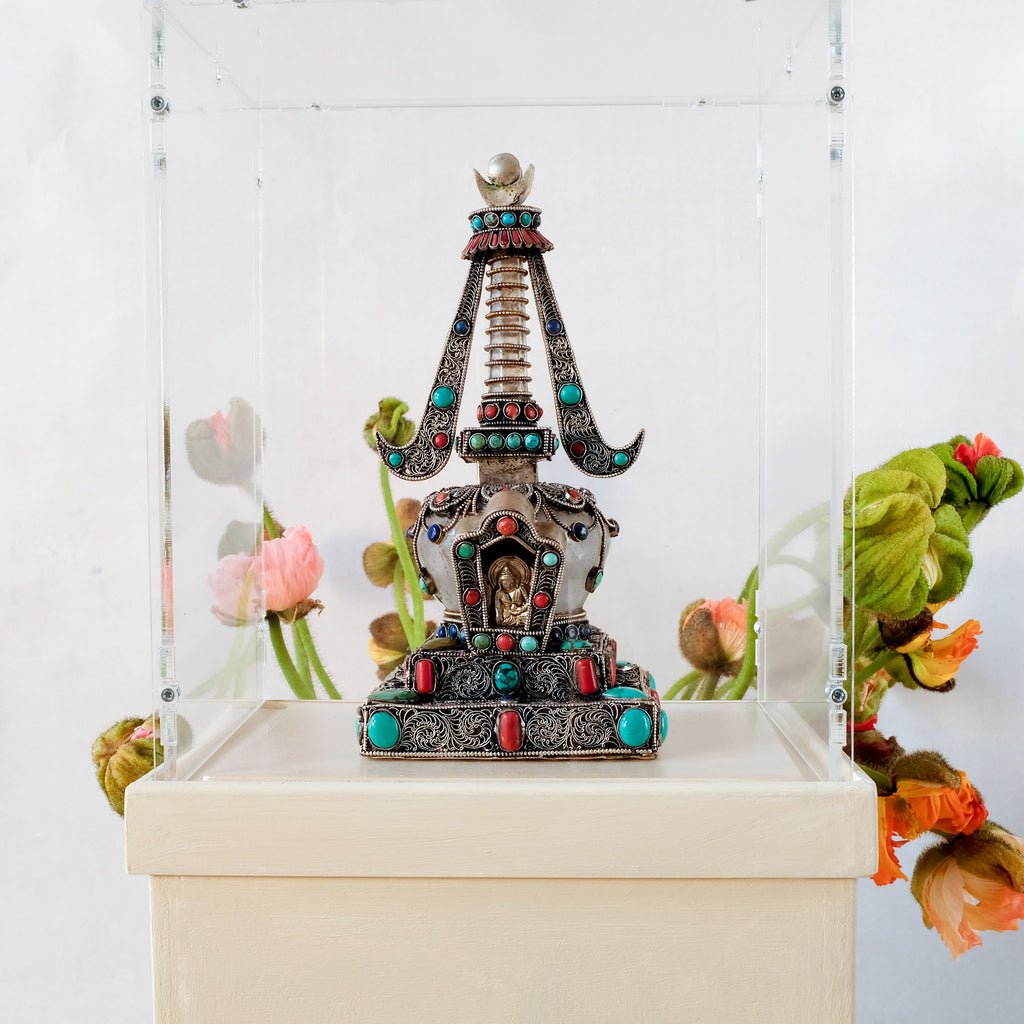 Stupa in einer Ausstellung, auf einem Sockel mit Acrylglashaube. Die Stupa hat einen Kristalldom und -mast. Sie ist mit Filigran gestaltet, versilbert und mit farbigen Steinen besetzt.