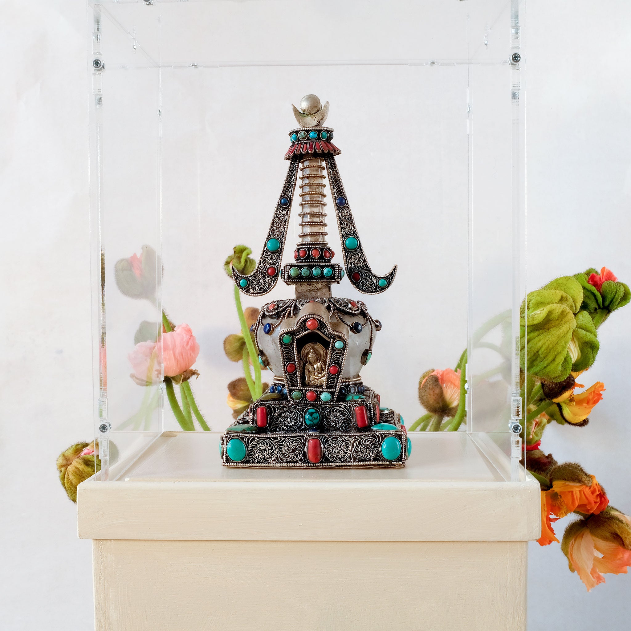 Stupa in einer Ausstellung, auf einem Sockel mit Acrylglashaube. Die Stupa hat einen Kristalldom und -mast. Sie ist mit Filigran gestaltet, versilbert und mit farbigen Steinen besetzt.