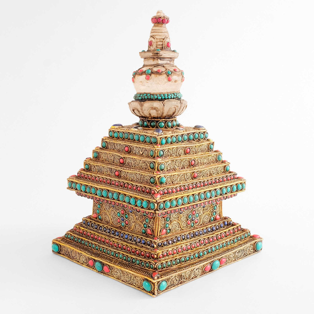 Stupa oder Chorten-Skulptur von einer Ecke aus gesehen. Die Stufen sind mit vergoldetem Perldraht umrahmt. Im Inneren dieses Rahmen sind feine Filigran-Spiralen zu Mustern angeordneten.