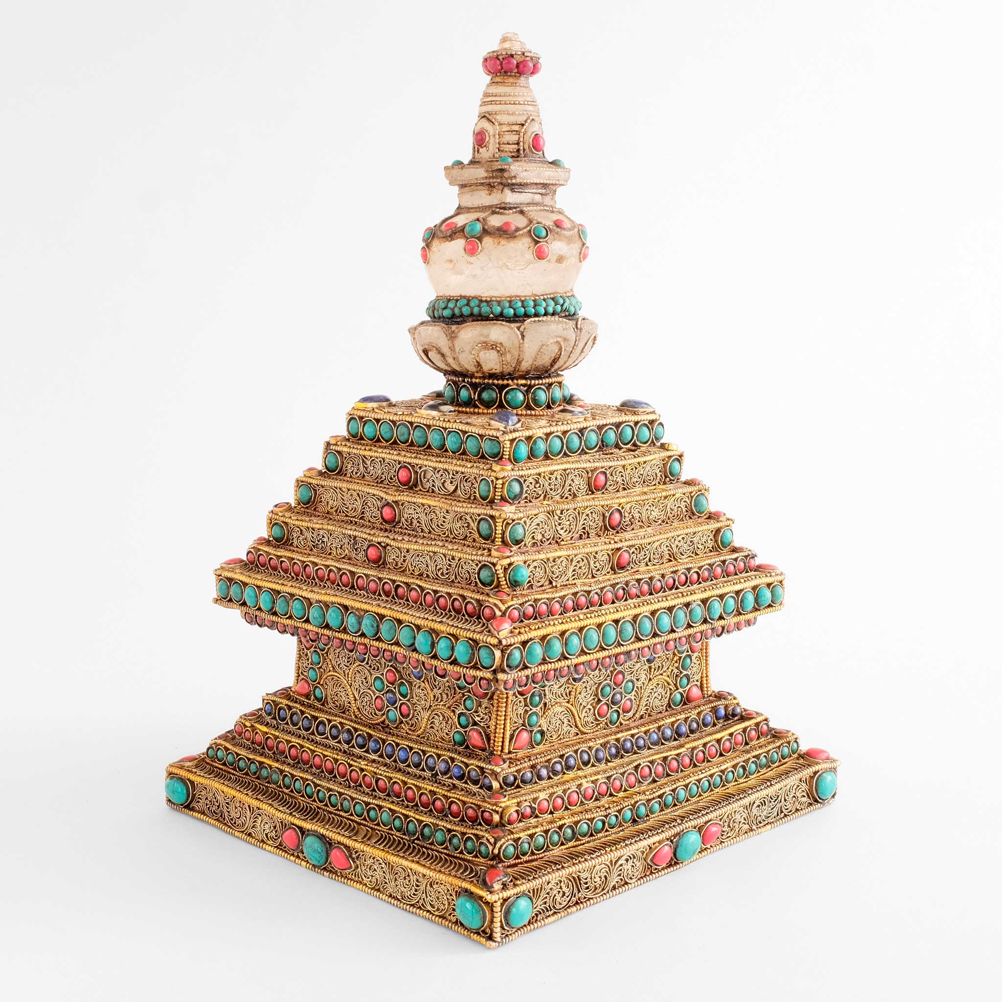 Stupa oder Chorten-Skulptur von einer Ecke aus gesehen. Die Stufen sind mit vergoldetem Perldraht umrahmt. Im Inneren dieses Rahmen sind feine Filigran-Spiralen zu Mustern angeordneten.