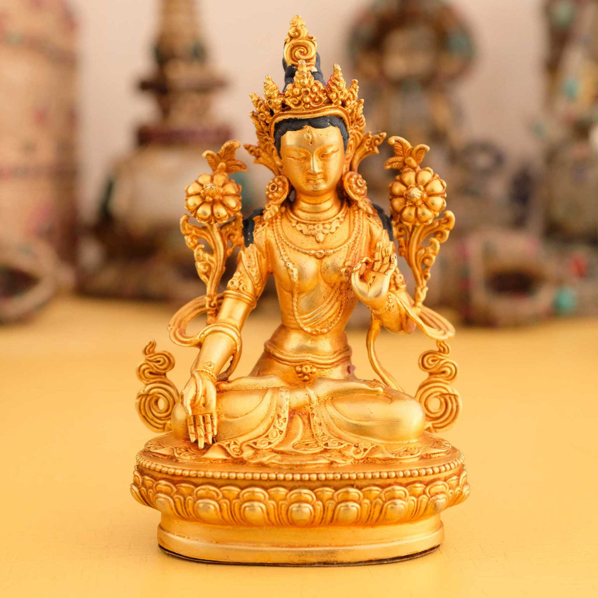 Kleine vergoldete Weiße Tara Statue, im Hintergrund rituelle Gegenstände des Vajrayana-Buddhismus in einer sanft beleuchteten Werkstattumgebung.